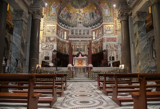 A Catholic priest is being filmed while conducting mass in front of empty pews. A woman standing far off to the side watches.