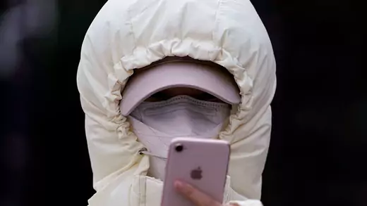 A woman wearing a mask and white jacket checks her pink iphone