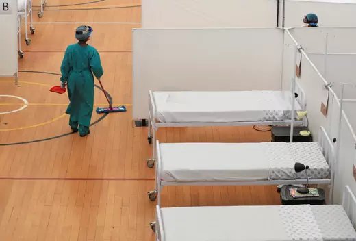 A health worker, in full personal protective equipment (PPE), mops the floor in a sports gym turned into a temporary coronavirus treatment facility. There are three freshly-made beds in the picture.