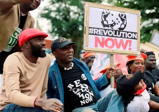 Several Nigerian people are seen sitting for a protest, mostly wearing red and black. Human rights activist Omoyele Sowore is seen holding a sign that reads "#RevolutionNOW!" in red and black text. His shirt uses the same slogan.