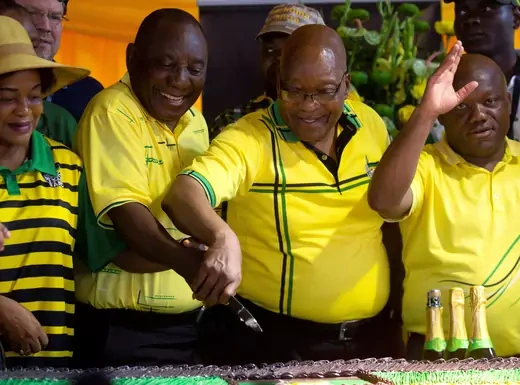 South African President Cyril Ramaphosa and former President Jacob Zuma, both wearing yellow shirts with black and green stripes, cut a cake together. Several others stand around them wearing shirts of the same coloring.