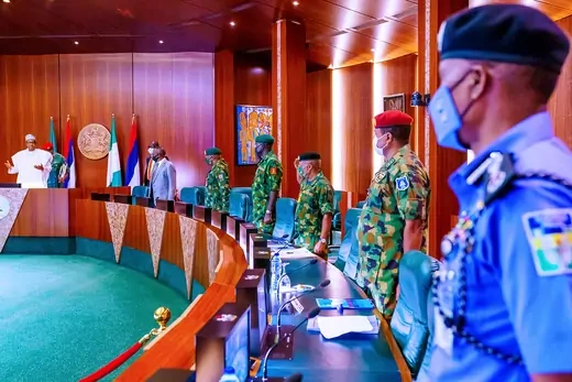 A group of men in military uniforms stand at attention around a long desk as Nigerian President Muhammadu Buhari gives an address.