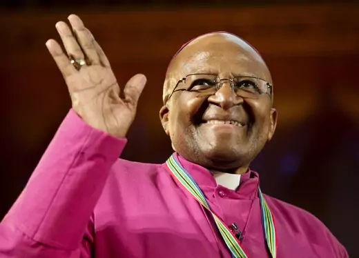 Desmond Tutu smiles and waves while wearing a red clerical tunic and collar.
