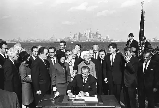 President Lyndon B. Johnson, surrounded by crowd, is seen signing the 1965 Immigration Act at the foot of the Statue of Liberty.