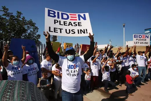 A migrant is seen holding up a sign saying "Biden please let us in" during a protest.