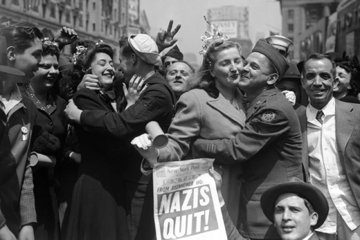 New Yorkers celebrate the surrender of Nazi Germany in Times Square, holding up a newspaper front page reading "Nazis Quit!"