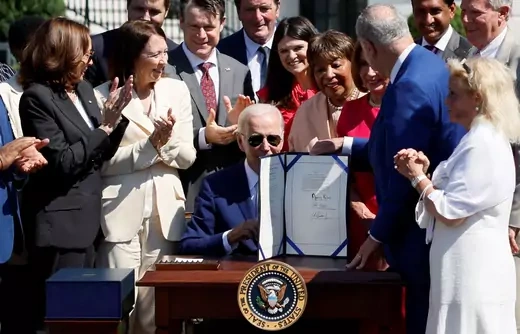 Joe Biden sits behind a desk with the Presidential Seal on it, flanked by a group of people clapping.