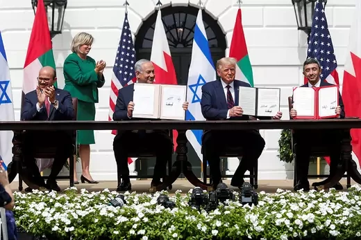 Four men in suits sitting at a long table, with three of them holding up signed agreements.  The other man and a woman behind them are clapping.  Flags of the United States, Bahrain, Israel, and the United Arab Emirates are in the background.