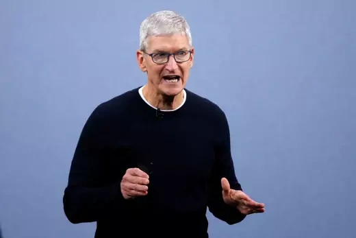 Tim Cook stands in a black sweater in front of a light blue background with his mouth partly open.