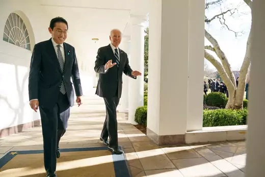 Kishida and Biden walk side by side in dark suits on a sunny outdoor colonnade. Both are smiling, and Biden is gesturing with his hands as he speaks.