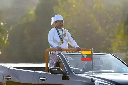 Myanmar's military leader stands in an open-top motorcade wearing traditional dress.