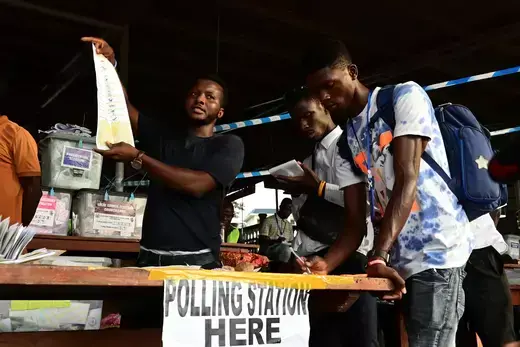 Election workers count ballots at a polling station in Sierra leone. A sign reads "polling station here." 