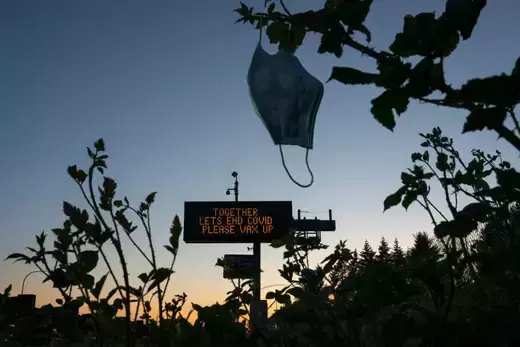 A highway sign promotes vaccination in the background. A discarded mask hangs off a branch in the foreground.