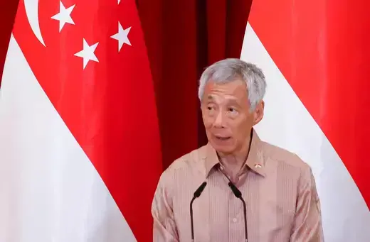 Prime Minister of Singapore stands in front of two white and red Singaporean flags while wearing a button-down shirt.