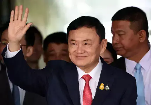Thai politician wears a navy suit and red tie while waving his hand.