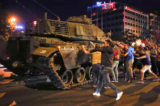 A group of men approach and bang with their hands a military tank in a downtown street at night.