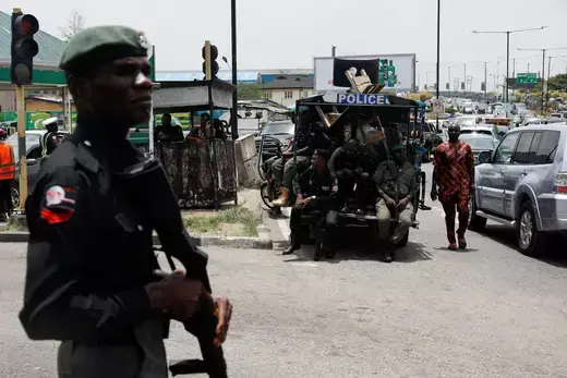 A Nigerian police officer is shown holding a rifle while a large police vehicle is shown in the background.