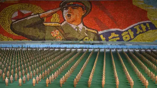 Army of soldiers dressed in solid beige uniforms standing in lines below a mural of a large military leader saluting.