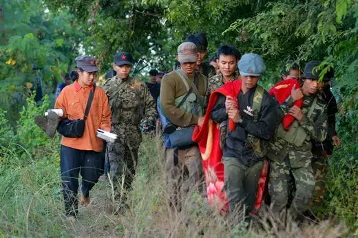Soldiers dressed in fatigues and dark hats walk through a forested area carrying a red blanket.