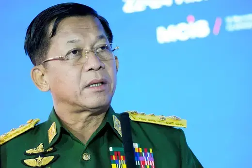 Myanmar general in green military uniform with gold medals wears glasses while speaking in front of a bright blue screen.