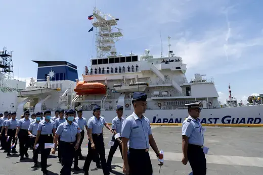 Coast guard officers in blue shirts and navy pants walk past a coast guard vessel on a dock.
