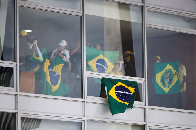 Supporters of Brazil's former President Jair Bolsonaro hold up Brazilian flags behind windows in government building.