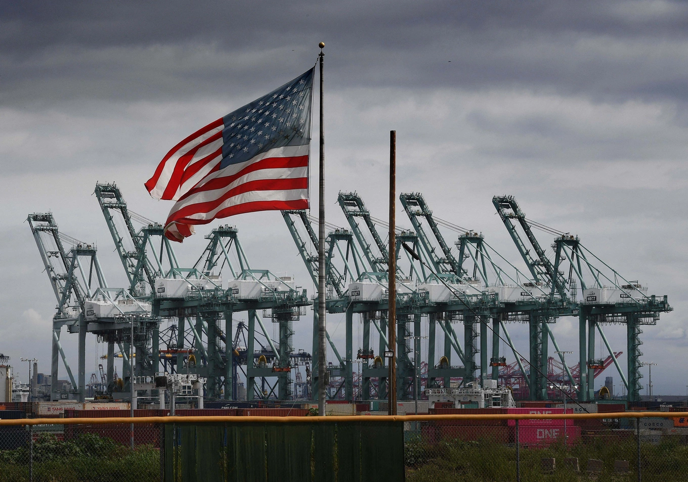 Picture of an American flag with sea port cranes in the background.