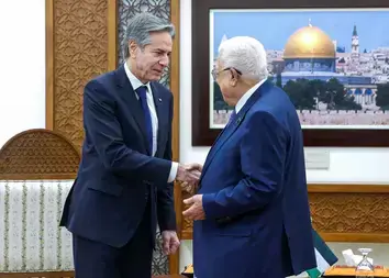 Two men in suits shake hands with a picture of Jerusalem on the wall and a Palestinian flag in the background. 