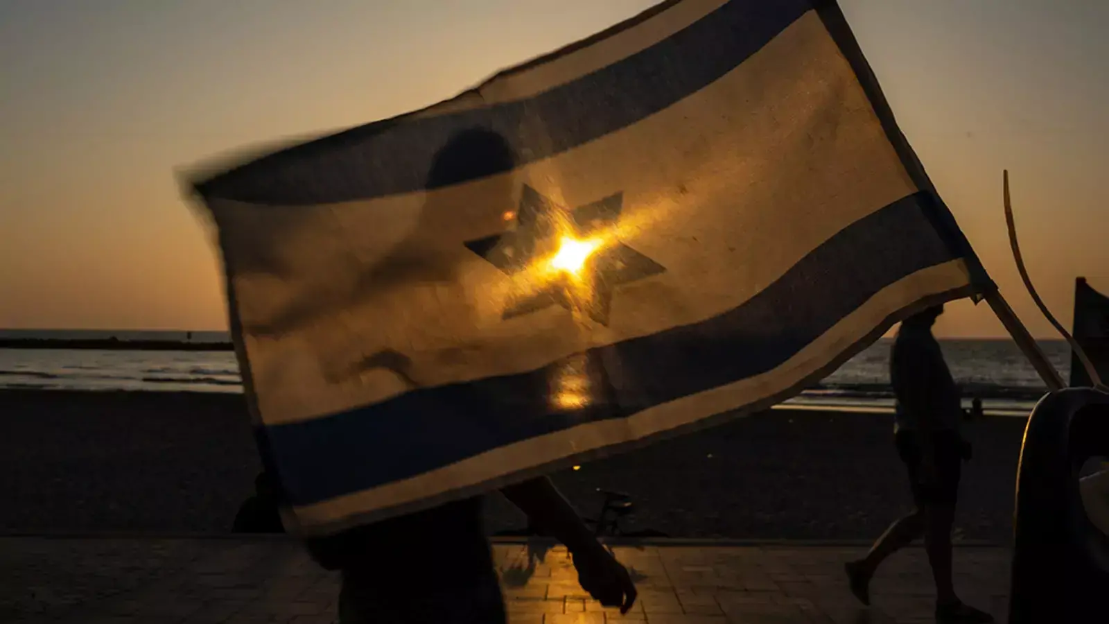 People walk past a waving Israeli flag as the sun on May 25, 2023 in Tel Aviv, Israel.