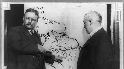 President Roosevelt pointing at a map of South America towards the area explored during the Roosevelt-Rondon Scientific Expedition in Brazil as another man looks on.