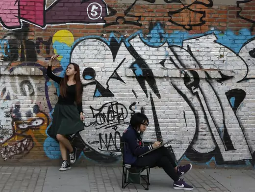 A woman poses for her self-portrait with her mobile phone while another works on a painting in front of a graffiti during the 2013 Beijing 798 Art Festival at the 798 Art Zone in Beijing October 8, 2013. The art zone, originally an unused factory, was transformed into a landmark of contemporary art in Beijing in the 1990s.