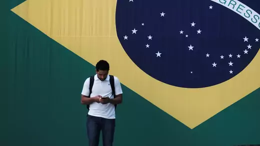 A man checks his mobile phone as a woman riding a bike passes next to a big Brazilian flag in Sao Paulo, Brazil June 28, 2018. 