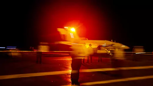 A blurry photo taken at night shows a military plane taking off from the deck of an American aircraft carrier as people watch.