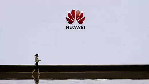 A woman in business clothing walks in front of a large screen that displays the red Huawei logo. 