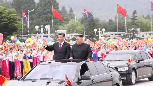 Xi Jinping and Kim Jong-un wear black suits, stand out the sunroof of a car, and wave to people in Pyongyang, North Korea.