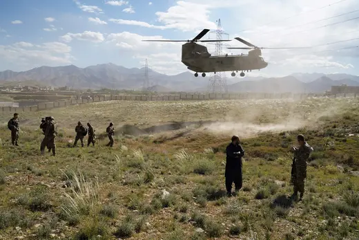 A military helicopter touches down in a barren field in Afghanistan. Several service members stand nearby. 