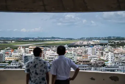 Two people look out from a balcony down on the Futenma air base. 