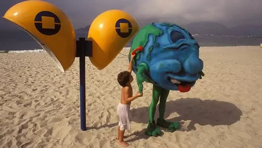 A boy holds up a red phone to a cartoon globe character on a beach. 