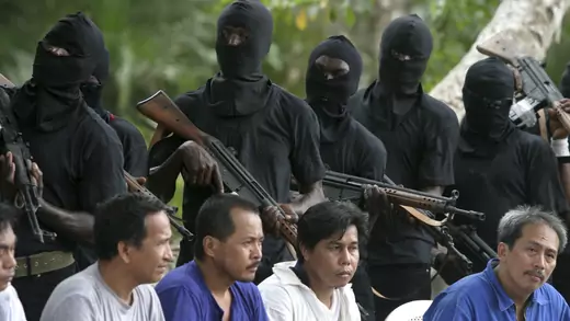 Filipino men sit and are guarded by masked militants carrying automatic weapons.