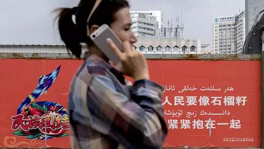 A young Uygur girl makes a phone call with her iPhone while walking past a propaganda wall.