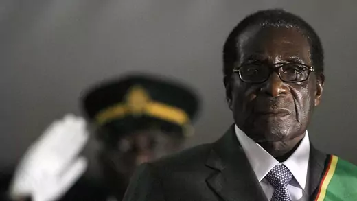 Robert Mugabe stands in front of a blurred out, saluting soldier.