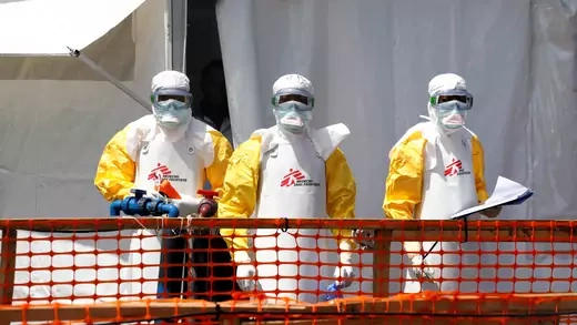 Health workers dressed in protective suits stand on the other side of an orange fence.