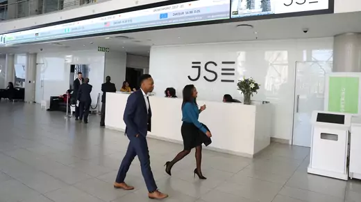 Visitors walk past a reception with an electronic board displaying movements in major indices at the Johannesburg Stock Exchange building