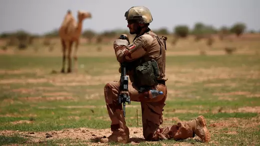 A French soldier kneels with his weapon while a camel stands in the background in a semi-arid landscape.