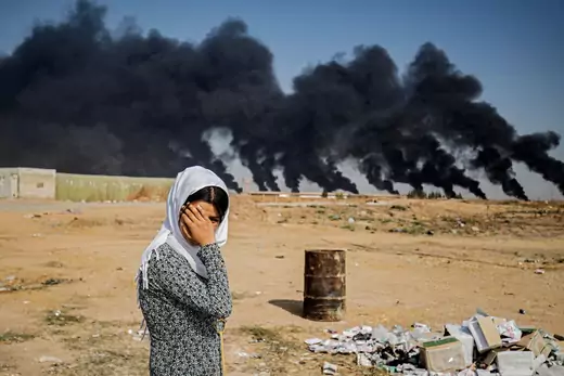 A woman covers her face as she stands on the outskirts of a town near the Syria-Turkey border, while smoke plumes meant to decrease visibility for Turkish warplanes billow from tire fires.