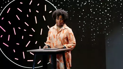 Nigerian writer Chimamanda Ngozi Adichie speaks at a podium, effectively a high-table. She is wearing a blouse of varying shades of orange, and is standing in front of a black background speckled with white.