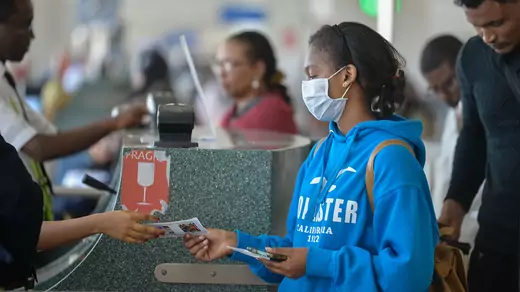 A young female traveller in a blue hoodie with a medical mask passes a ticket to the gate agent at the Addis airport.