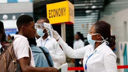 A female health worker in a white shirt, medical gloves, and a blue mask, uses an infrared thermometer to check the temperature of a young, male traveller who  as part of the coronavirus screening procedure at the Kotoka International Airport in Accra, Ghana, on January 30, 2020. Behind them are other passengers and health workers and a yellow "Economy" sign.