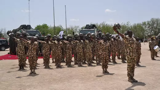 Nigerian soldiers in military fatigues hold their helmets up and to the right in a salute for President Buhari (not pictured). Behind them are two armored military vehicles and a red carpet.