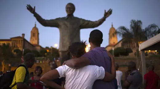 A tall man in a purple polo shirt puts his arms around the shoulders of a shorter woman in a white shirt as she holds his shoulder. They both look at the statue of Nelson Mandela, raising his hands and smiling, in a small crowd of people. The statue is flanked by two towers attached to building with roman columns.
